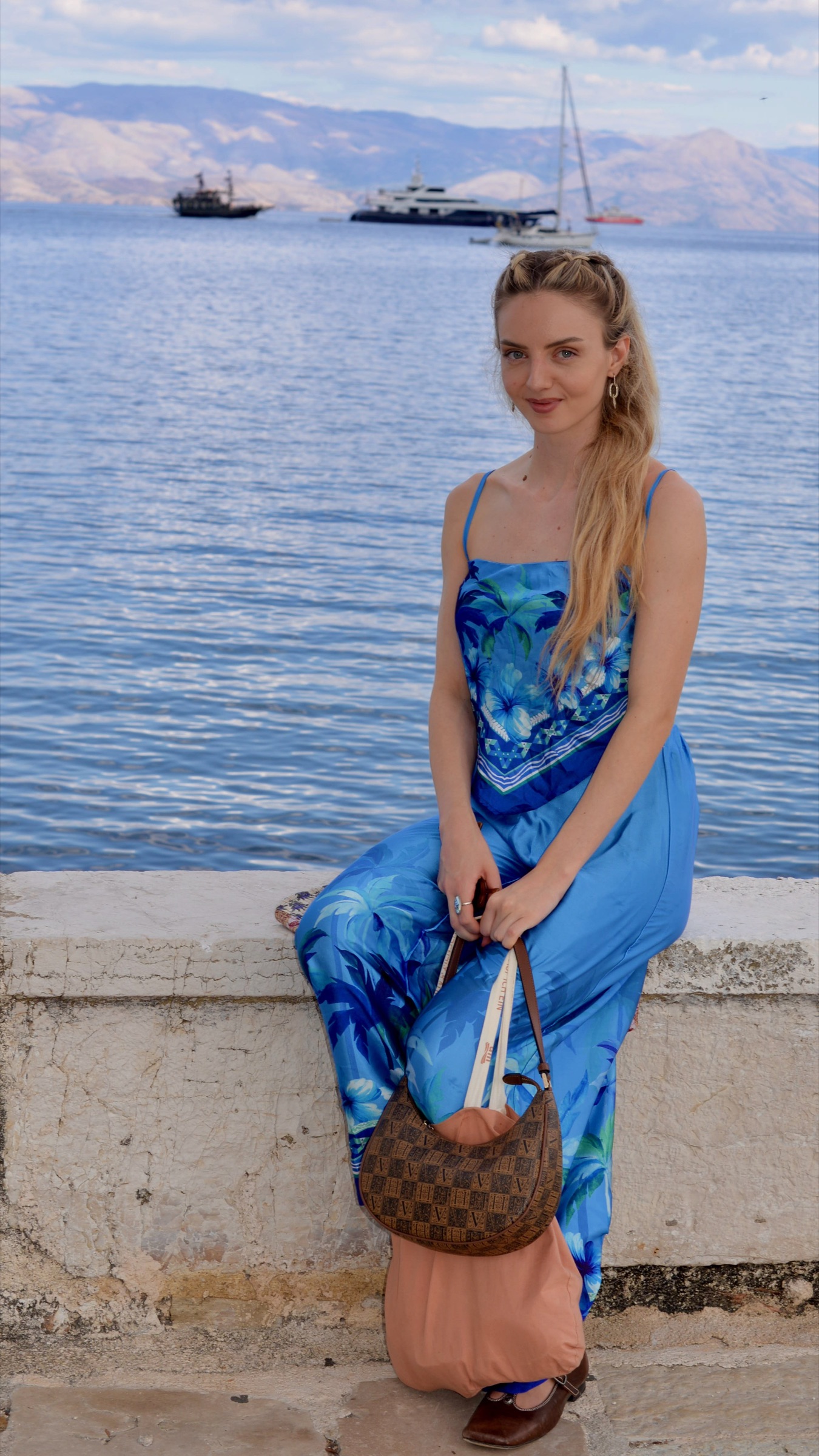 A woman in a blue dress sitting on the stone seawall in Corfu Town, boats and mainland mountains behind her