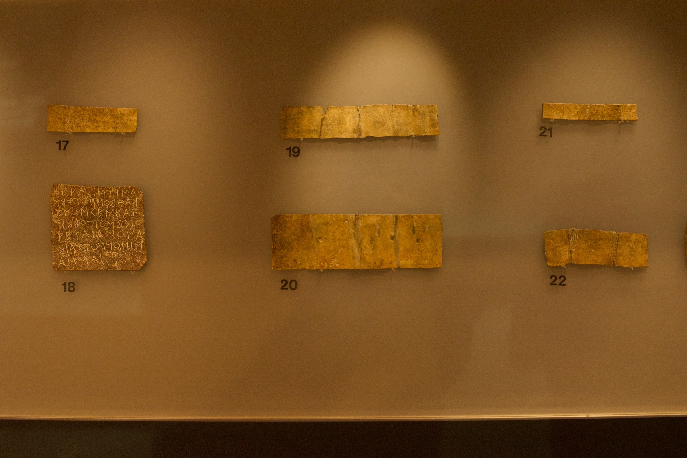 Small lead strips with scratched Greek letters in a museum display case, numbered 17 to 22