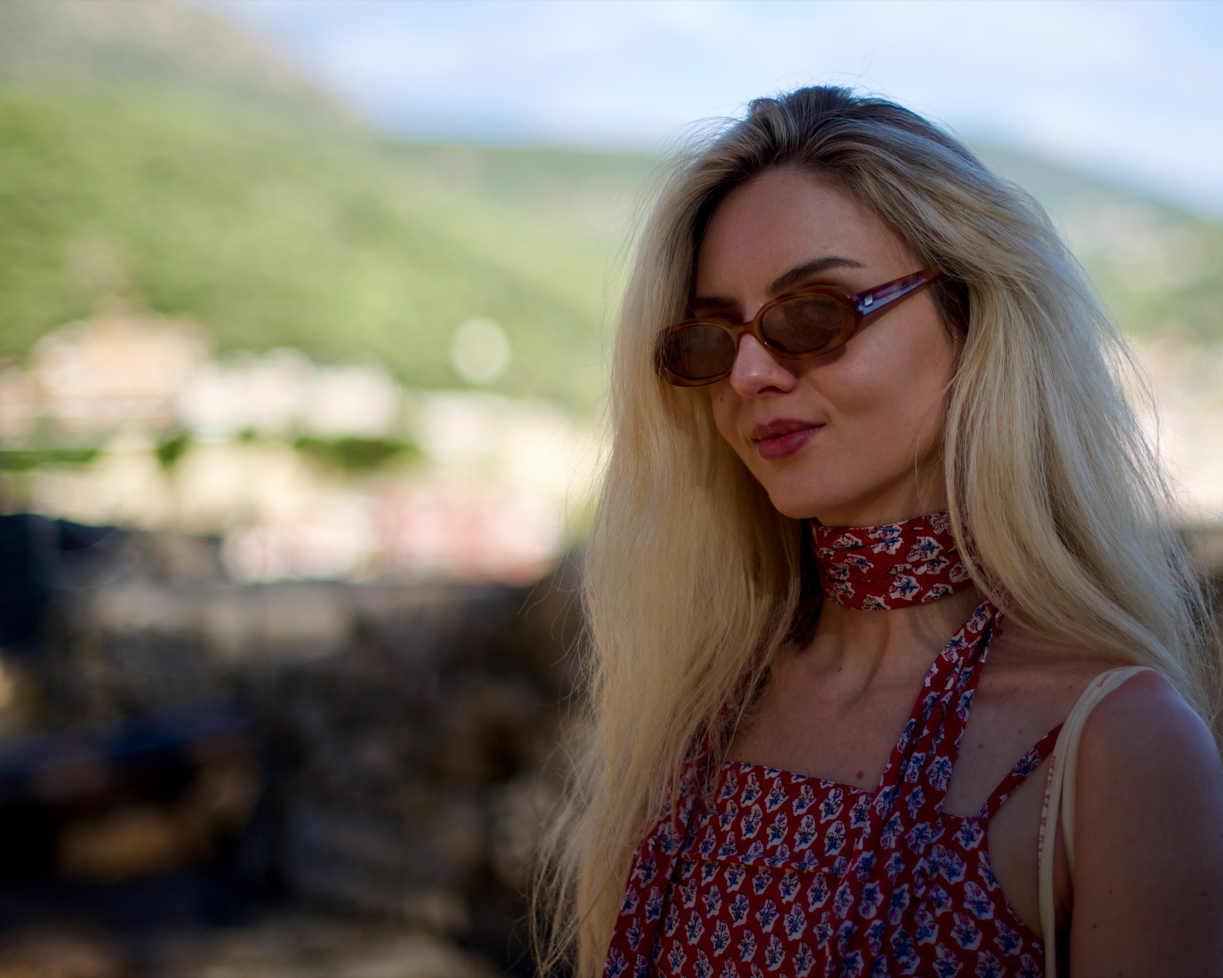 Portrait of a woman with sunglasses and a red scarf, the town of Parga blurred behind her